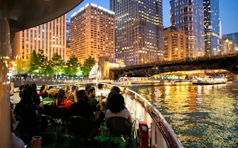Cruise passengers on Chicago River at night with city skyline and bridge in view.