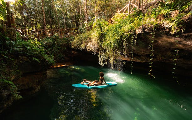 Couple relaxing on a paddleboard in a lush cenote surrounded by greenery.