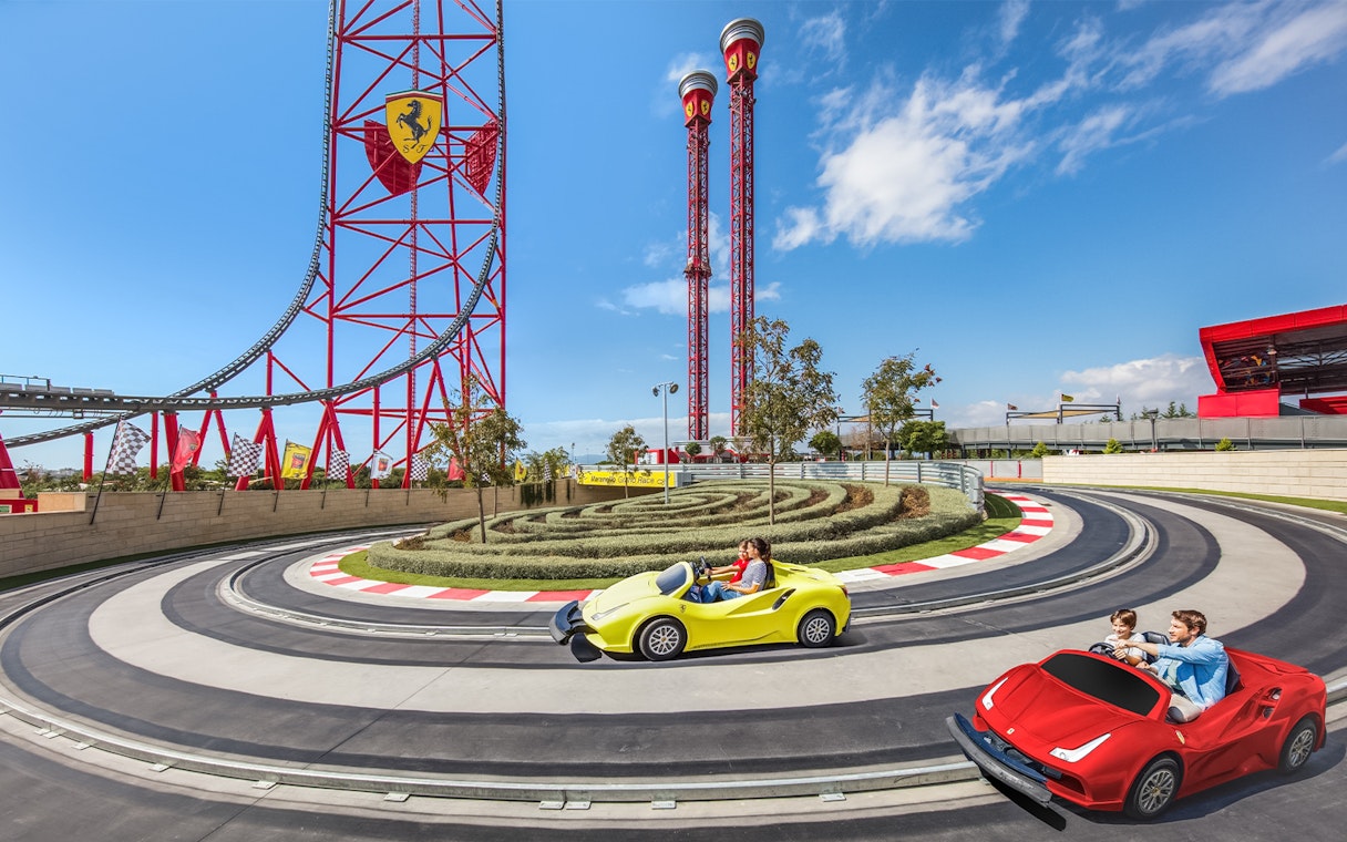 Visitors driving mini Ferraris on a race track at Ferrari Land, with roller coaster in background.