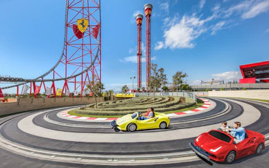 Visitors driving mini Ferraris on a race track at Ferrari Land, with roller coaster in background.