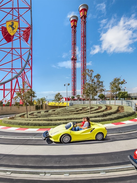 Visitors driving mini Ferraris on a race track at Ferrari Land, with roller coaster in background.