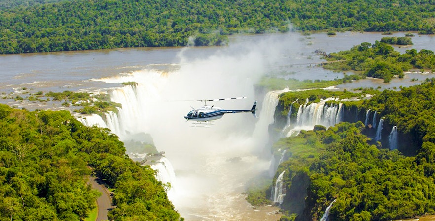 Helicopter flying over Iguazú Falls surrounded by lush greenery.