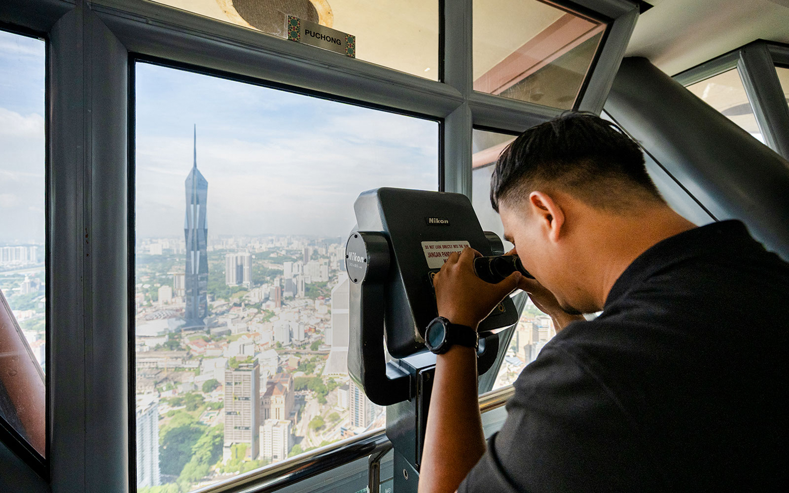 Tourist using binoculars at Kuala Lumpur Tower Observation Deck with city view.