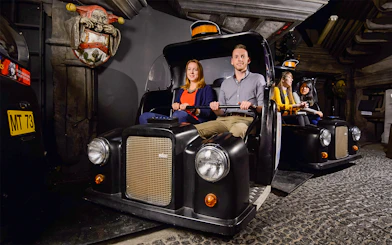 Visitors on Spirit of London Ride in black taxi-themed cars at an indoor attraction.