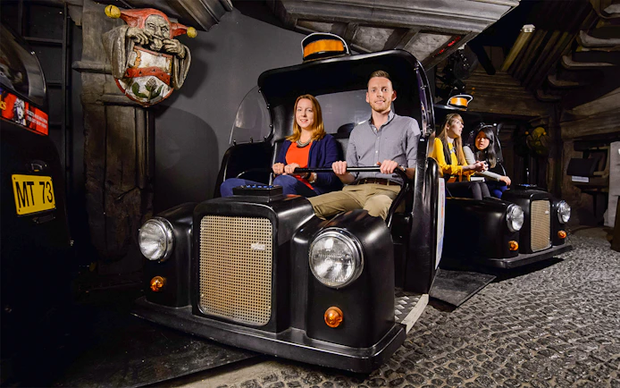 Visitors on Spirit of London Ride in black taxi-themed cars at an indoor attraction.