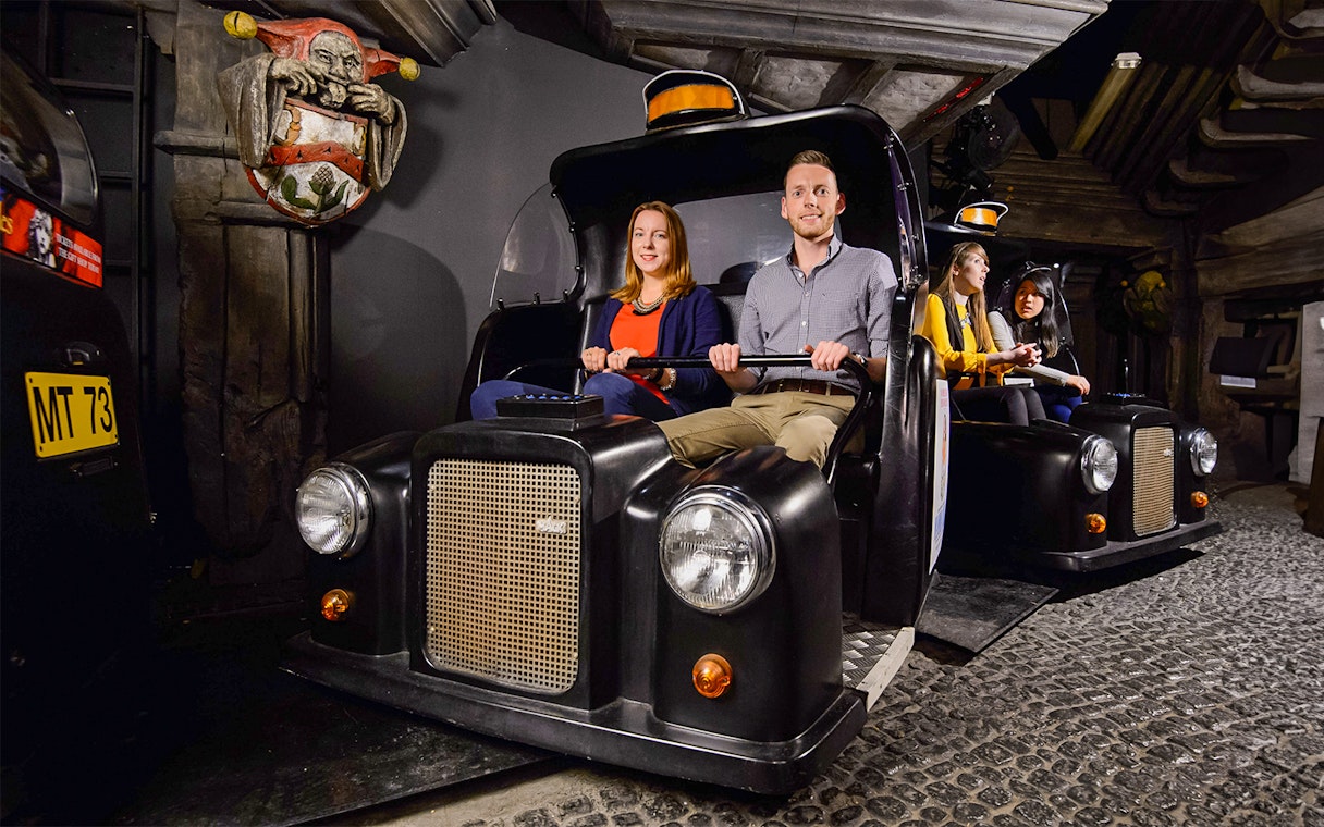 Visitors on Spirit of London Ride in black taxi-themed cars at an indoor attraction.