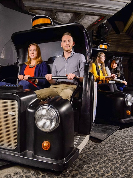 Visitors on Spirit of London Ride in black taxi-themed cars at an indoor attraction.