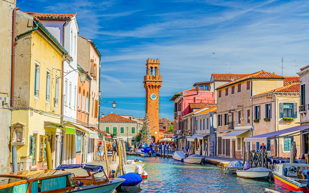 Murano island canal with boats and clock tower in the background, Venice, Italy.