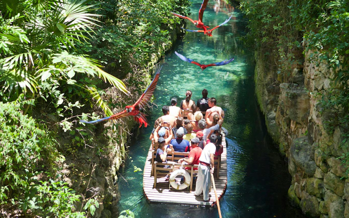 Tourists on a boat ride through a lush canal with macaws flying overhead at Xcaret Park.