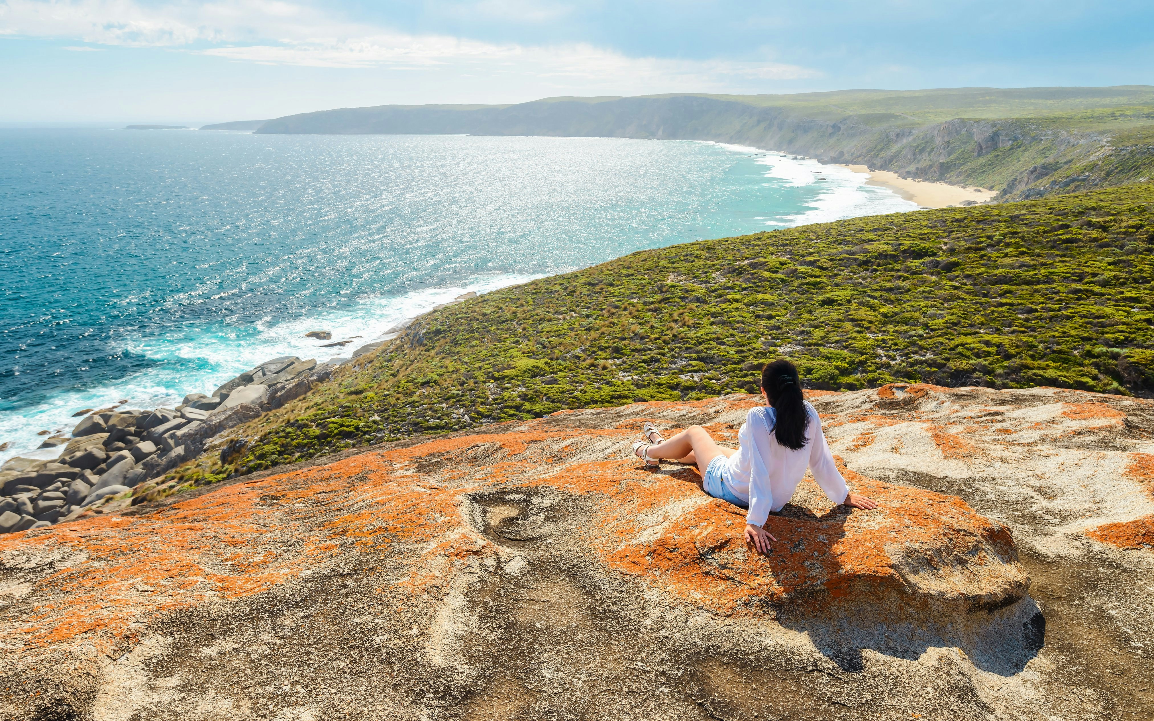 Woman sitting on Remarkable Rocks cliff, overlooking ocean and coastline, Kangaroo Island.