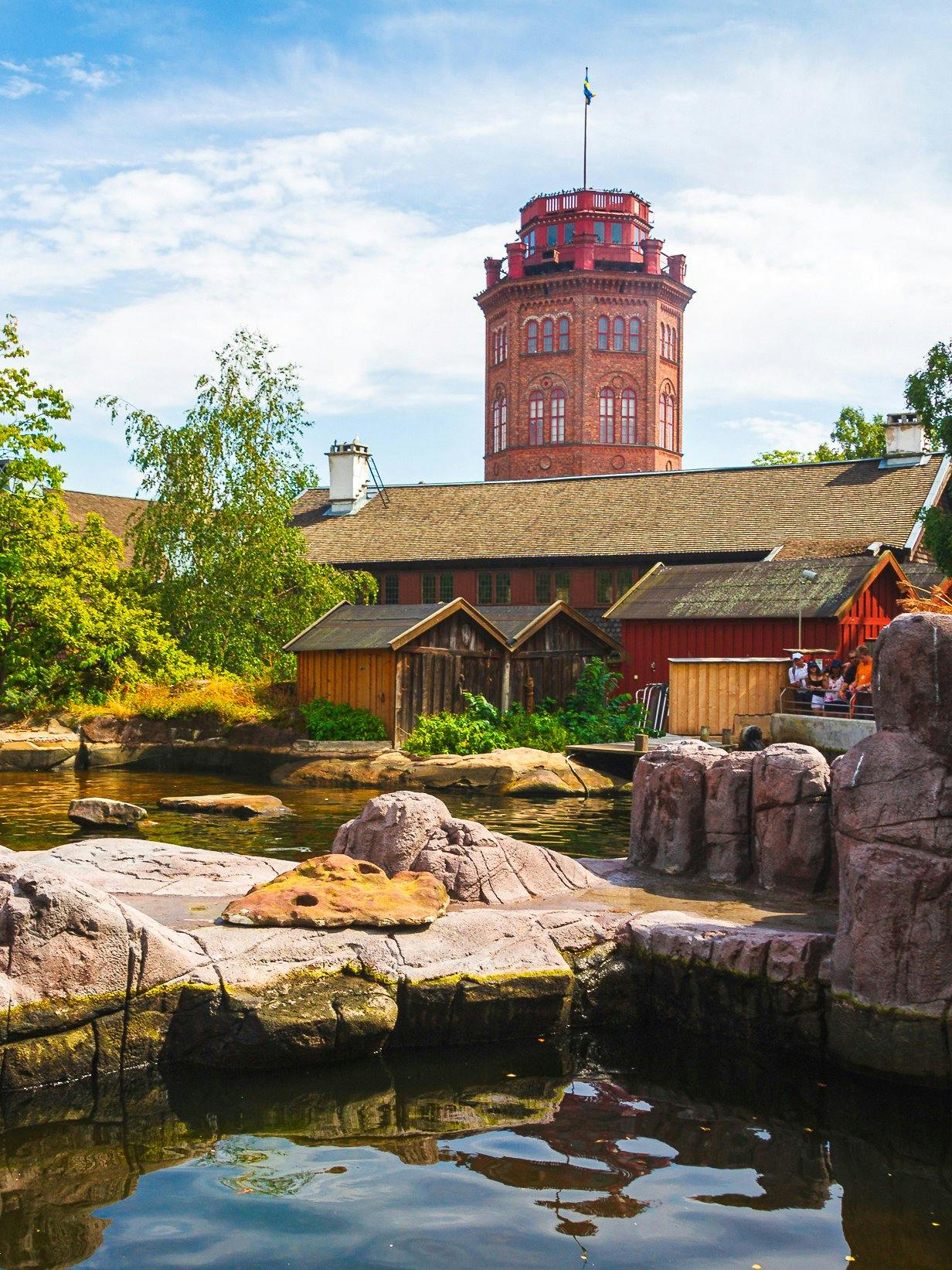 Skansen Museum tower with surrounding traditional wooden buildings and rocky water feature.