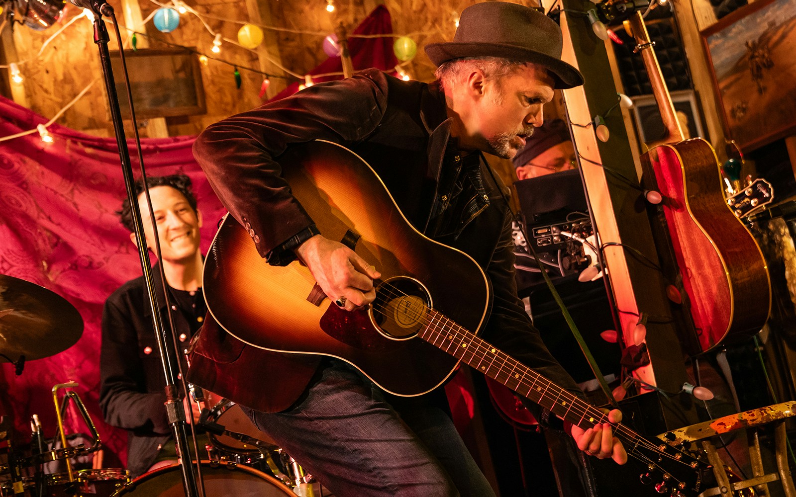 Musician playing guitar on stage with drummer, surrounded by colorful lights.