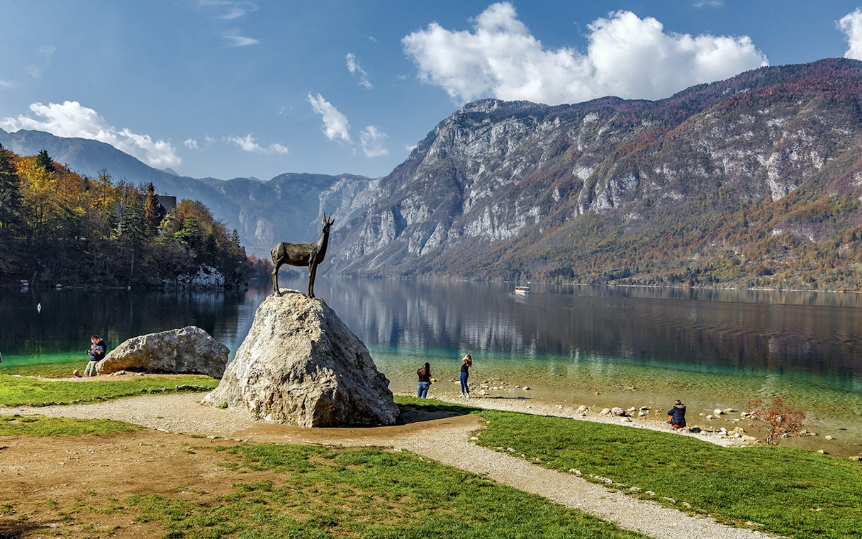 Guests by Bohinj Lake with a goat statue and mountain backdrop in Slovenia.