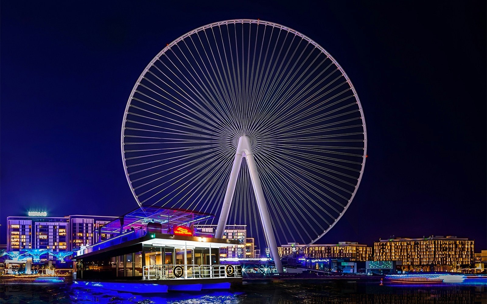 Ain Dubai Ferris wheel illuminated at night with a houseboat in the foreground.
