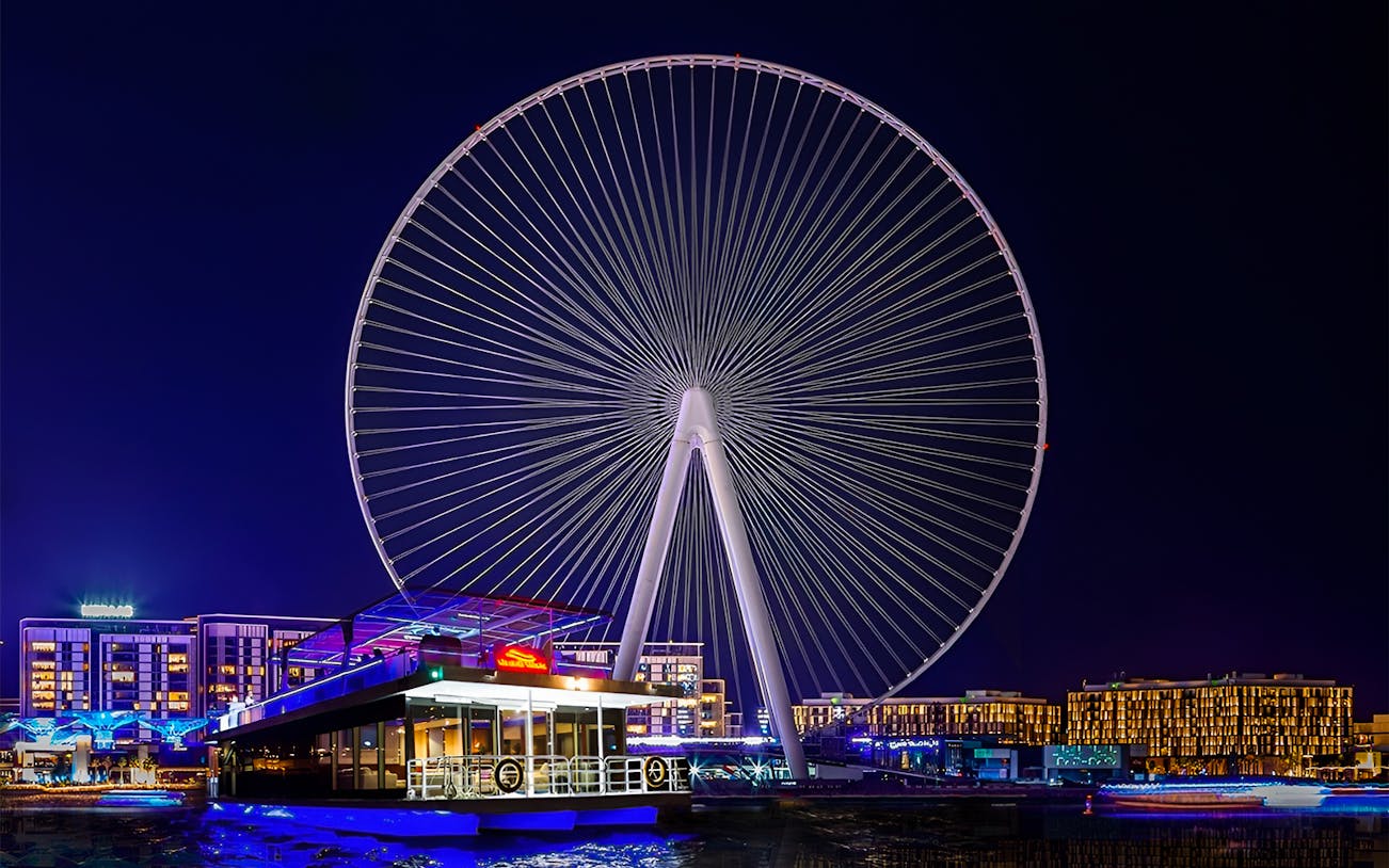 Ain Dubai Ferris wheel illuminated at night with a houseboat in the foreground.