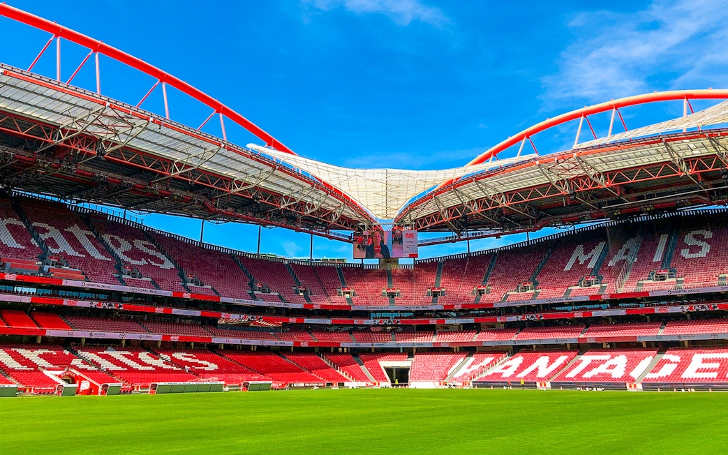 Benfica Stadium ground view with red seating and green field under blue sky.