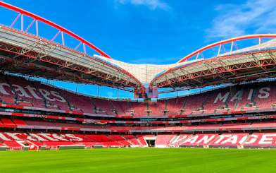 Benfica Stadium ground view with red seating and green field under blue sky.