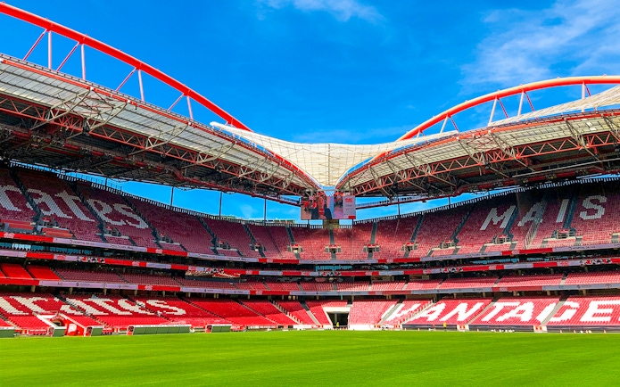 Benfica Stadium ground view with red seating and green field under blue sky.