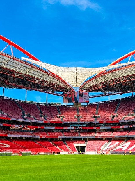 Benfica Stadium ground view with red seating and green field under blue sky.