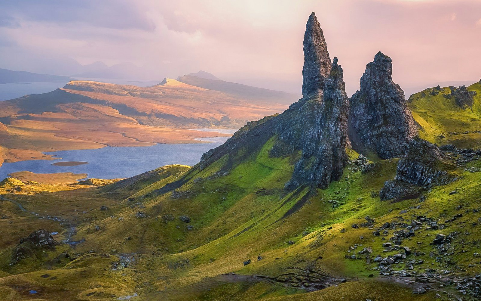 Rock formations and green hills in Glencoe, Scotland, with a distant view of a lake and mountains.