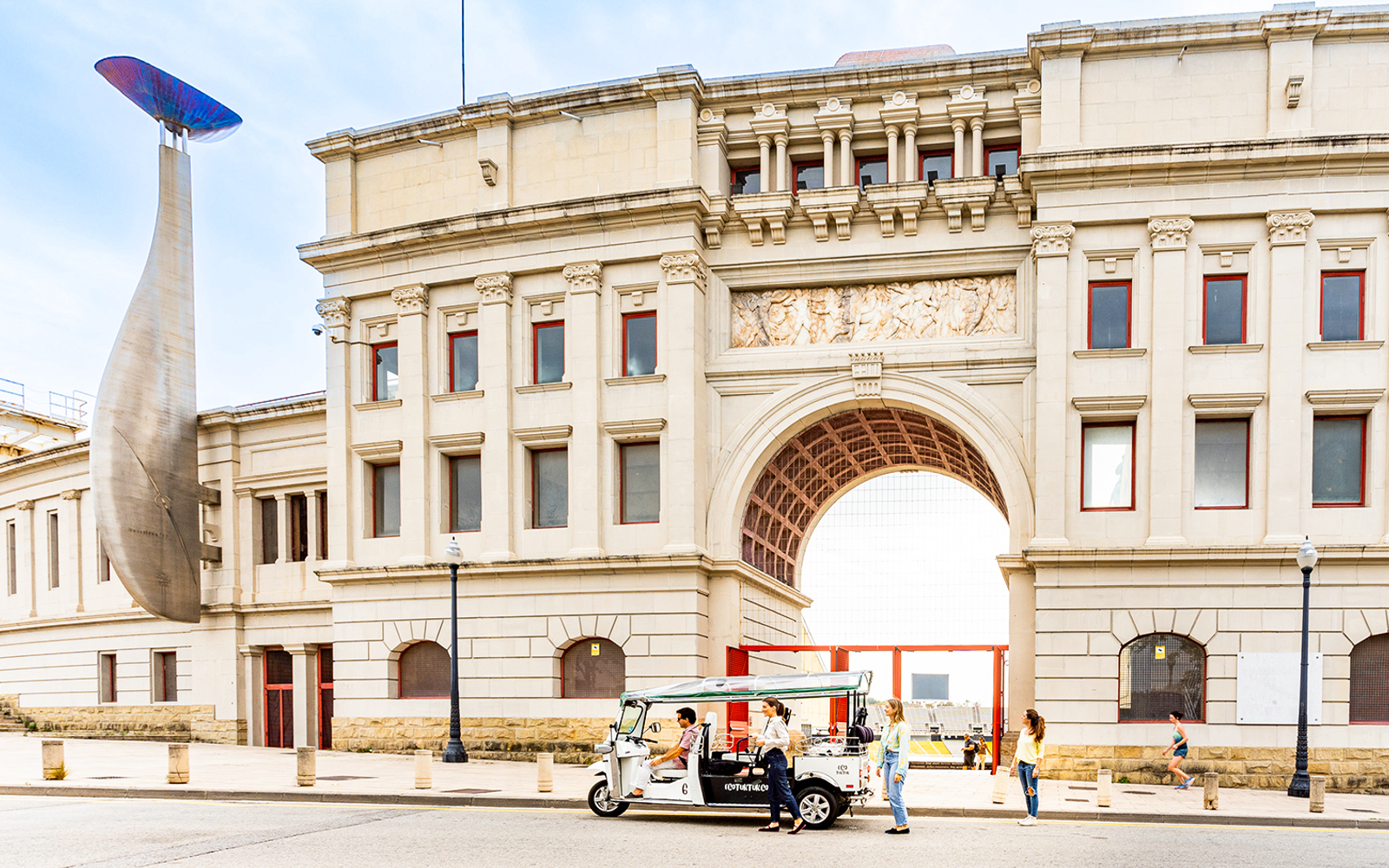 Tuk tuk tour in front of Barcelona's historic building with unique sculpture.