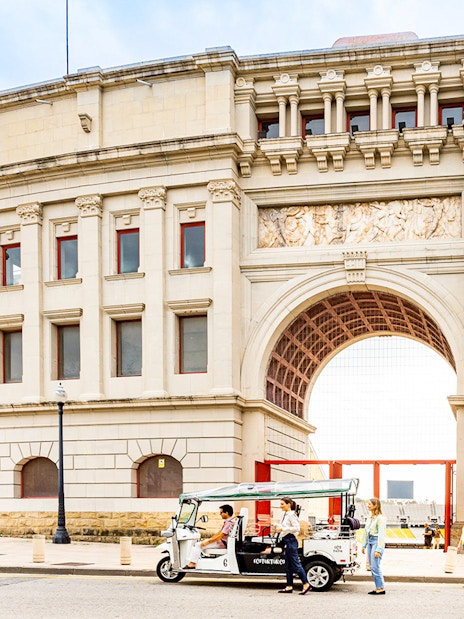 Tuk tuk tour in front of Barcelona's historic building with unique sculpture.