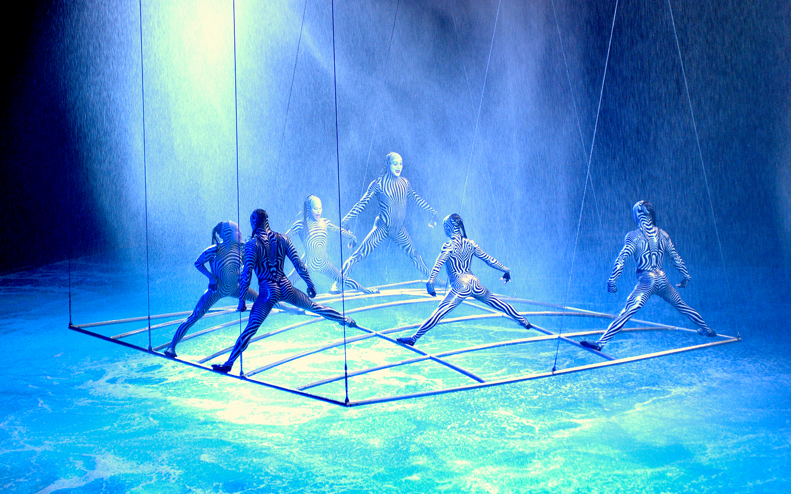 Performers in striped costumes on a suspended grid in a water-themed show.