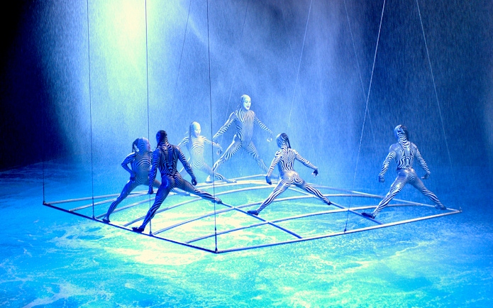 Performers in striped costumes on a suspended grid in a water-themed show.