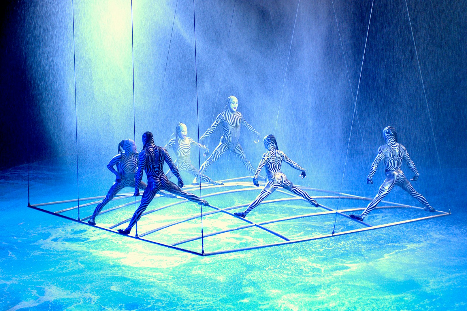 Performers in striped costumes on a suspended grid in a water-themed show.