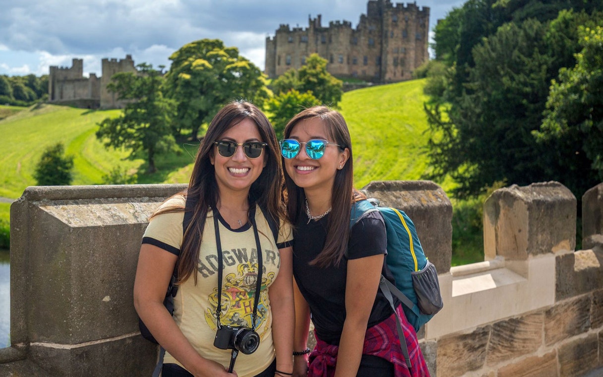 Two tourists smiling with Alnwick Castle in the background on a day tour from Edinburgh.