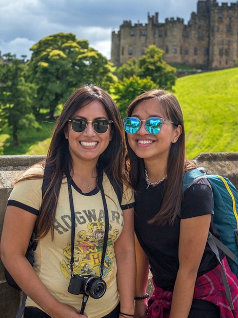 Two tourists smiling with Alnwick Castle in the background on a day tour from Edinburgh.