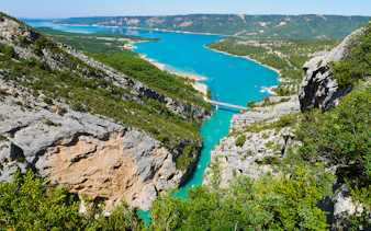 Gorges du Verdon with turquoise river and bridge, surrounded by cliffs and greenery.