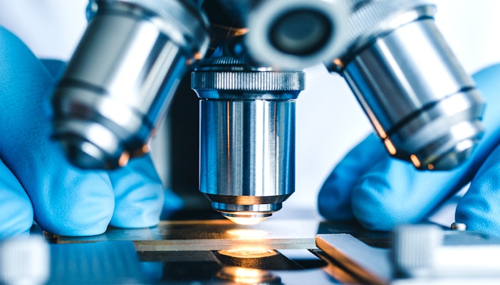 Microscope lens focused on a slide with gloved hands adjusting.