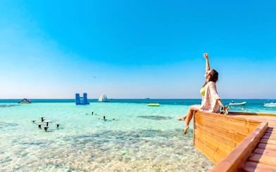 Woman relaxing on a wooden deck by clear waters at Orange Bay, Hurghada, with inflatable structures in the sea.
