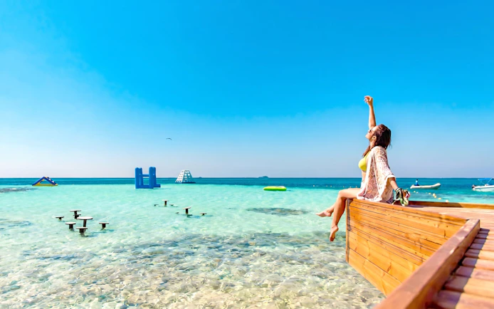Woman relaxing on a wooden deck by clear waters at Orange Bay, Hurghada, with inflatable structures in the sea.