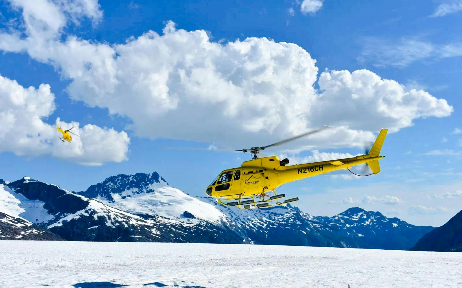 Yellow helicopter flying over snowy glacier in Juneau, Alaska, with mountain backdrop.