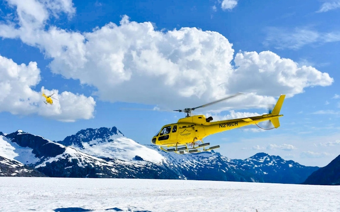Yellow helicopter flying over snowy glacier in Juneau, Alaska, with mountain backdrop.