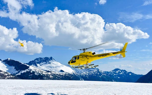 Yellow helicopter flying over snowy glacier in Juneau, Alaska, with mountain backdrop.