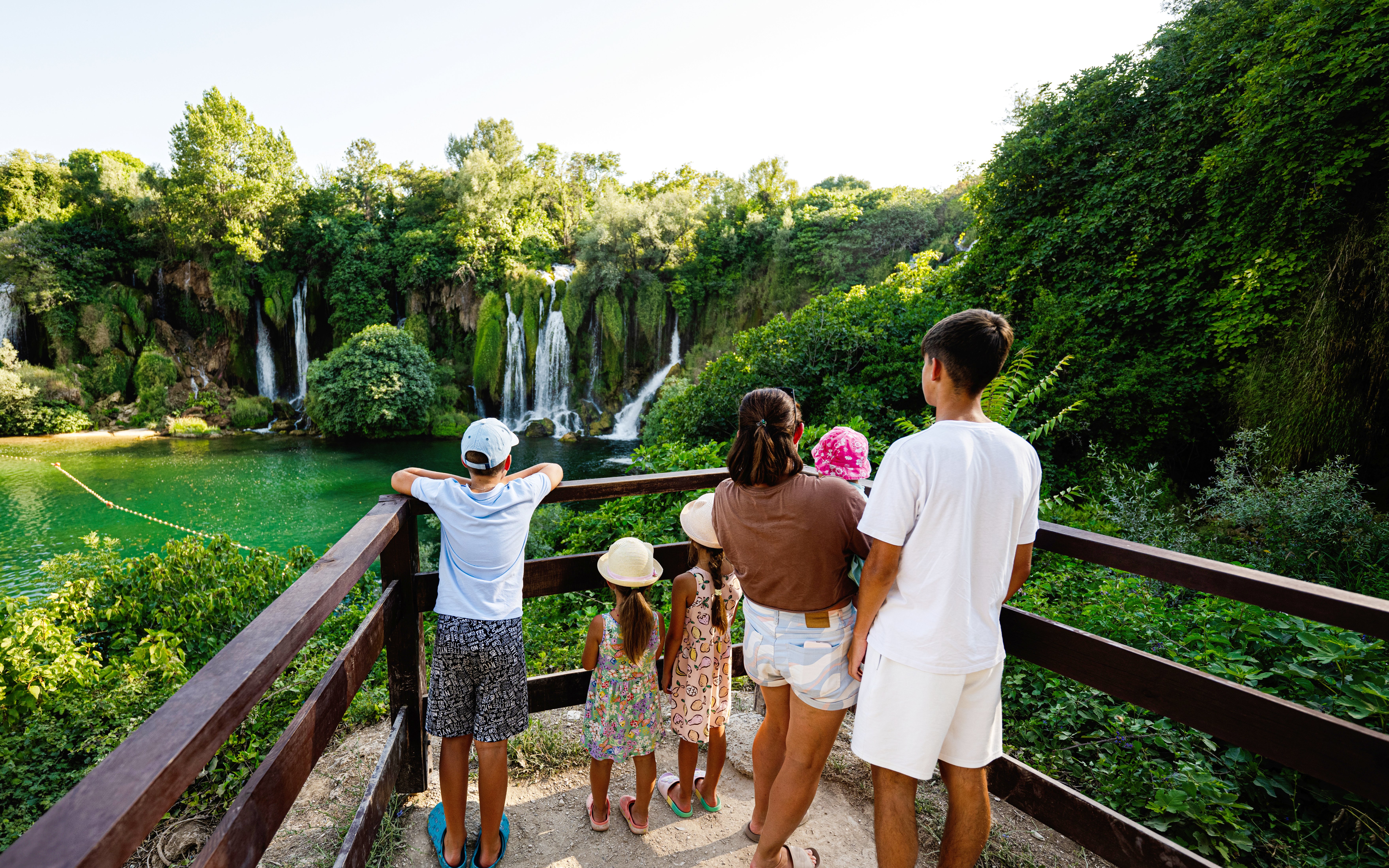 Family viewing Kravice Waterfall from a wooden platform surrounded by lush greenery.