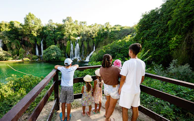Family viewing Kravice Waterfall from a wooden platform surrounded by lush greenery.