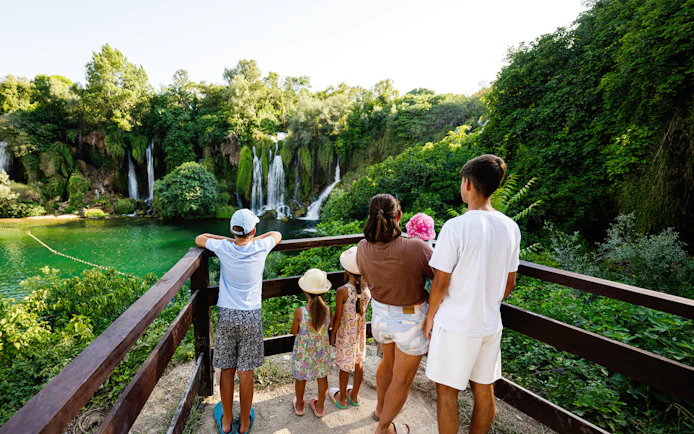 Family viewing Kravice Waterfall from a wooden platform surrounded by lush greenery.