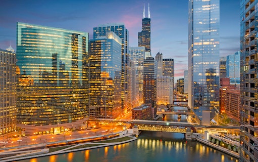 Chicago skyline with illuminated skyscrapers and the Chicago River at dusk.