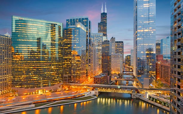 Chicago skyline with illuminated skyscrapers and the Chicago River at dusk.