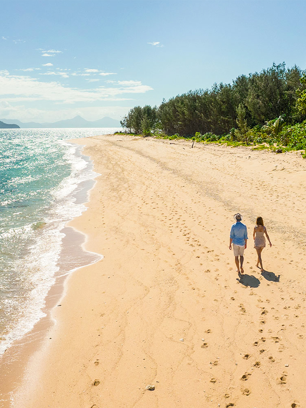 Couple walking on a sandy beach at Frankland Islands, with ocean and trees in the background.