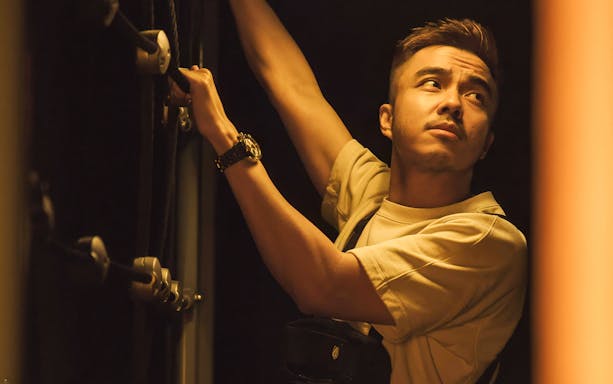 Man participating in Malaysia Breakout The Curve Spy Game, climbing a rope in dim lighting.
