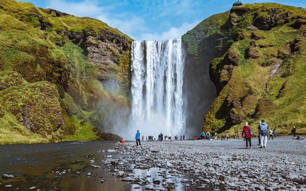 Visitors at Skogafoss waterfall in Iceland, surrounded by lush green cliffs.