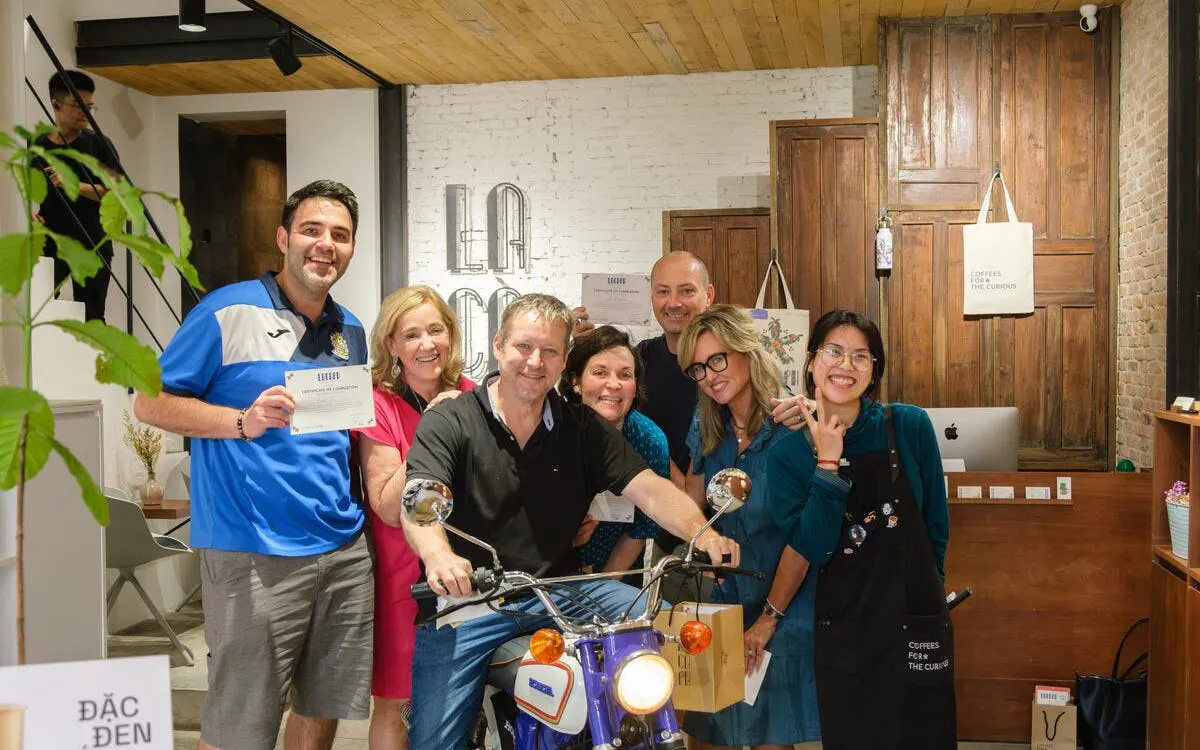 Group enjoying Vietnamese coffee workshop, posing with certificates and a vintage motorcycle.