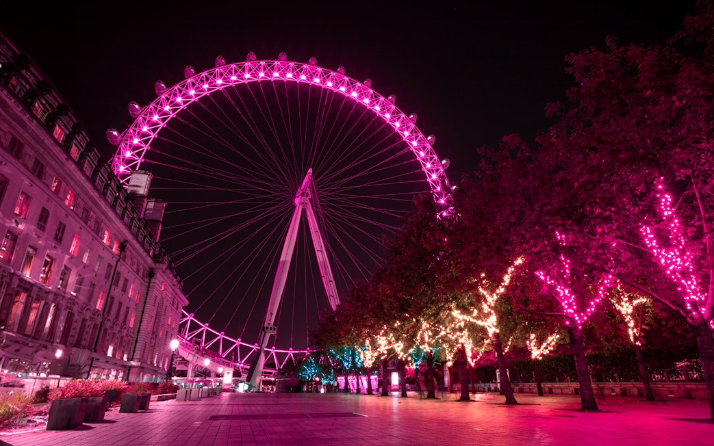 London Eye illuminated at night with vibrant pink lights, view from the riverside walkway.