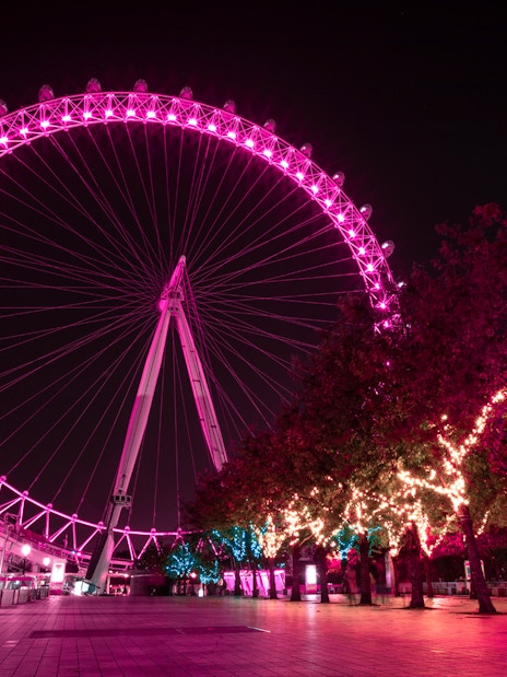 London Eye illuminated at night with vibrant pink lights, view from the riverside walkway.