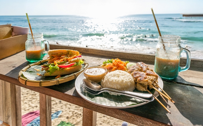 Balinese meal with rice, satay, and drinks on a beachside table in Nusa Penida, Bali.
